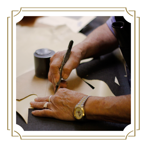 Close-up of hands wearing a wristwatch and a ring, cutting patterns out of leather with a utility knife on a workspace.